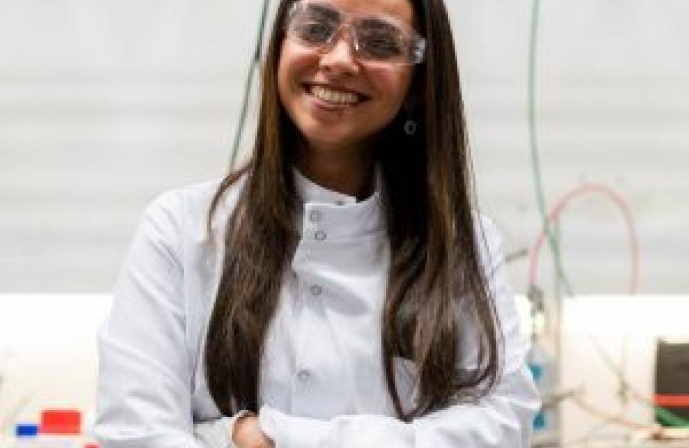 A young girl in a lab coat and safety classes standing in a lab space smiling.