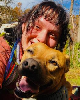 Woman with dark brown hair and bangs poses with their dog while hiking