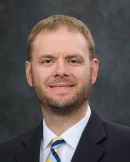 Portrait of Jake Sparks from the shoulders up, wearing a navy blue jacket, white shirt, and blue and gold striped tie.