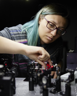 Lab photo of Jasmine Hinton aligning an optical system with red laser. In the photo she has blue hair and red glasses.