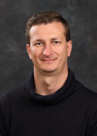 a smiling person in a black turtleneck in front of a grey portrait background